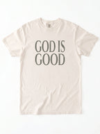 Cream T-shirt laid flat with distressed gray text reading “God is Good.”