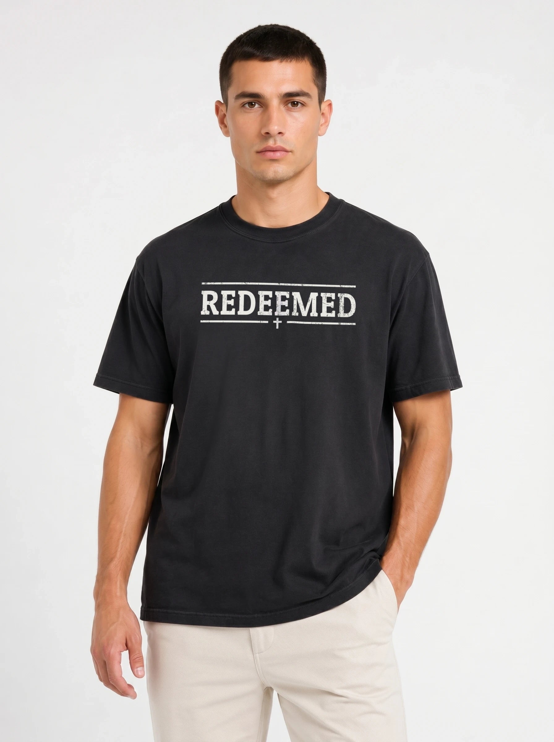 A man wearing a black short-sleeve t-shirt with the text 'REDEEMED' printed in distressed white block letters across the chest, flanked by two horizontal lines and a small cross below, paired with light beige pants, against a plain light gray background.