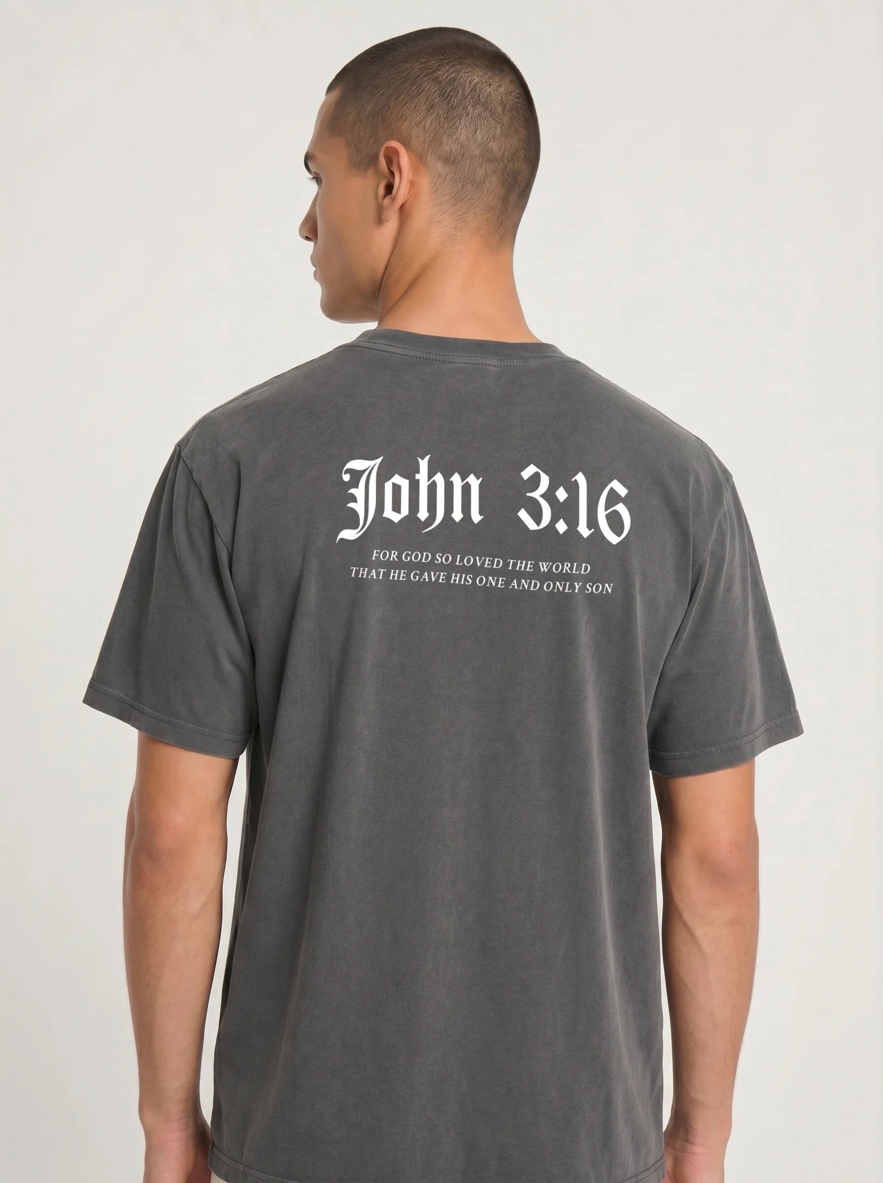 A man wearing a dark gray washed t-shirt, viewed from the back. The shirt features the text 'John 3:16' in a white Gothic font, with the subtitle 'FOR GOD WHO LOVED THE WORLD THAT HE GAVE HIS ONE AND ONLY SON' printed below in smaller white text.