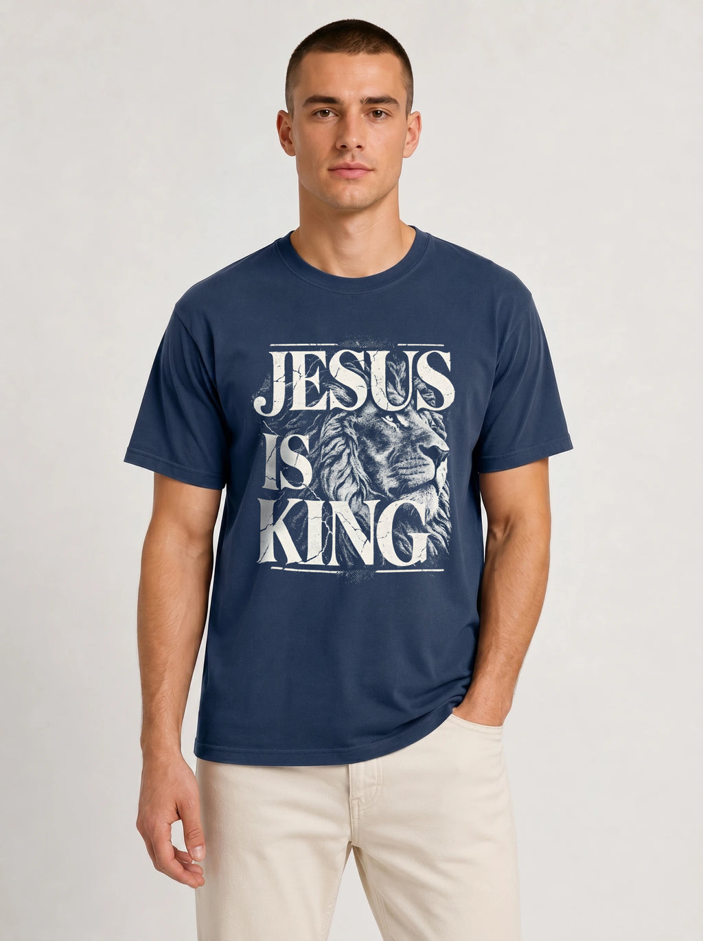 A man wearing a navy blue crewneck t-shirt with a distressed white graphic that reads 'JESUS IS KING' over an illustration of a lion's head, paired with light beige pants, against a light gray studio background.