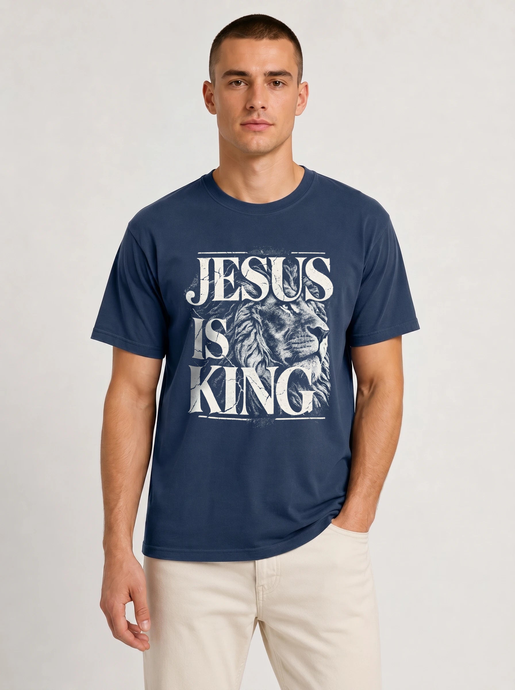 A man wearing a navy blue crewneck t-shirt with a distressed white graphic that reads 'JESUS IS KING' over an illustration of a lion's head, paired with light beige pants, against a light gray studio background.