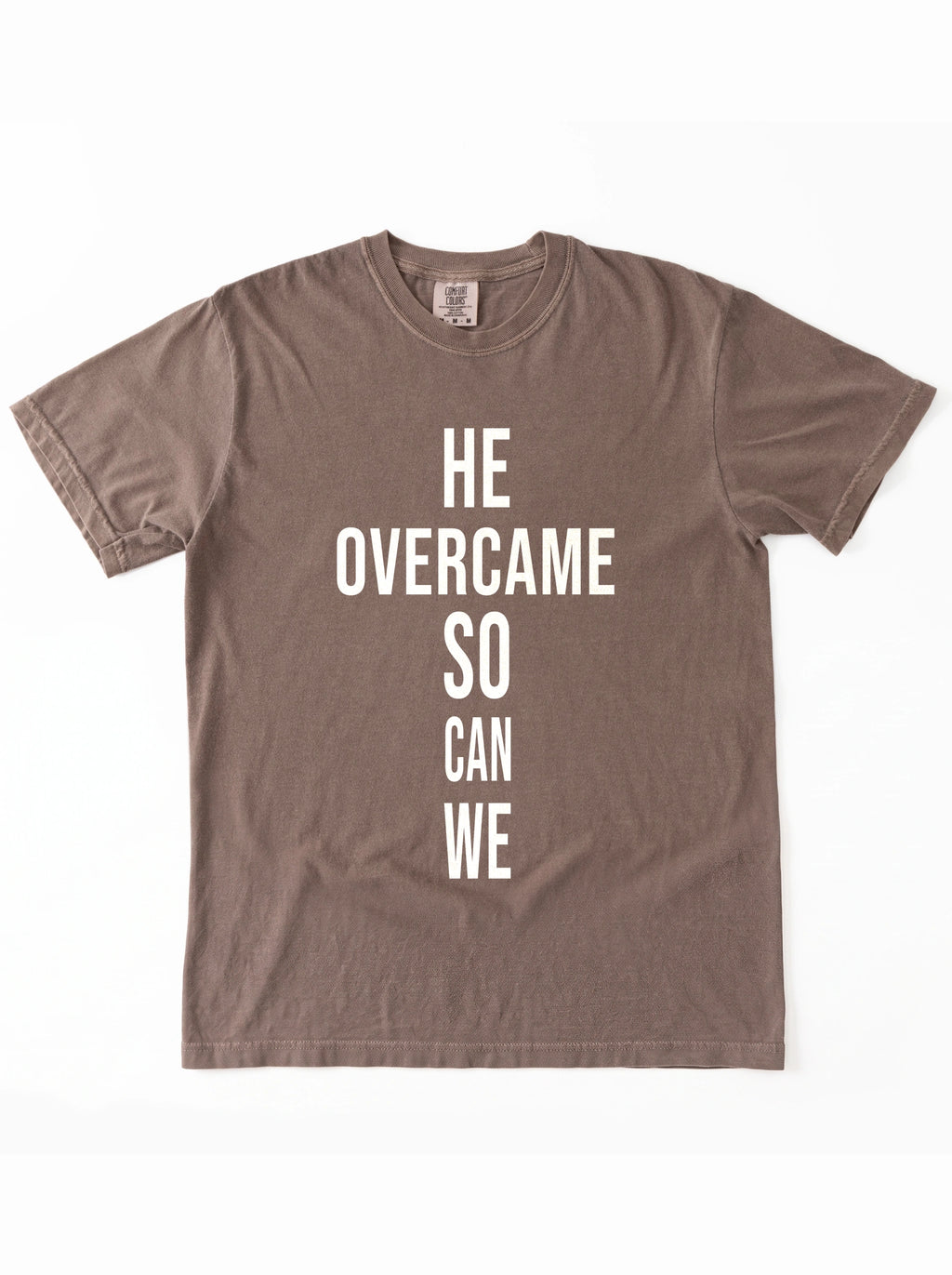 Faded brown T-shirt laid flat with large white text reading “HE OVERCAME SO CAN WE.”