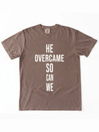 Faded brown T-shirt laid flat with large white text reading “HE OVERCAME SO CAN WE.”