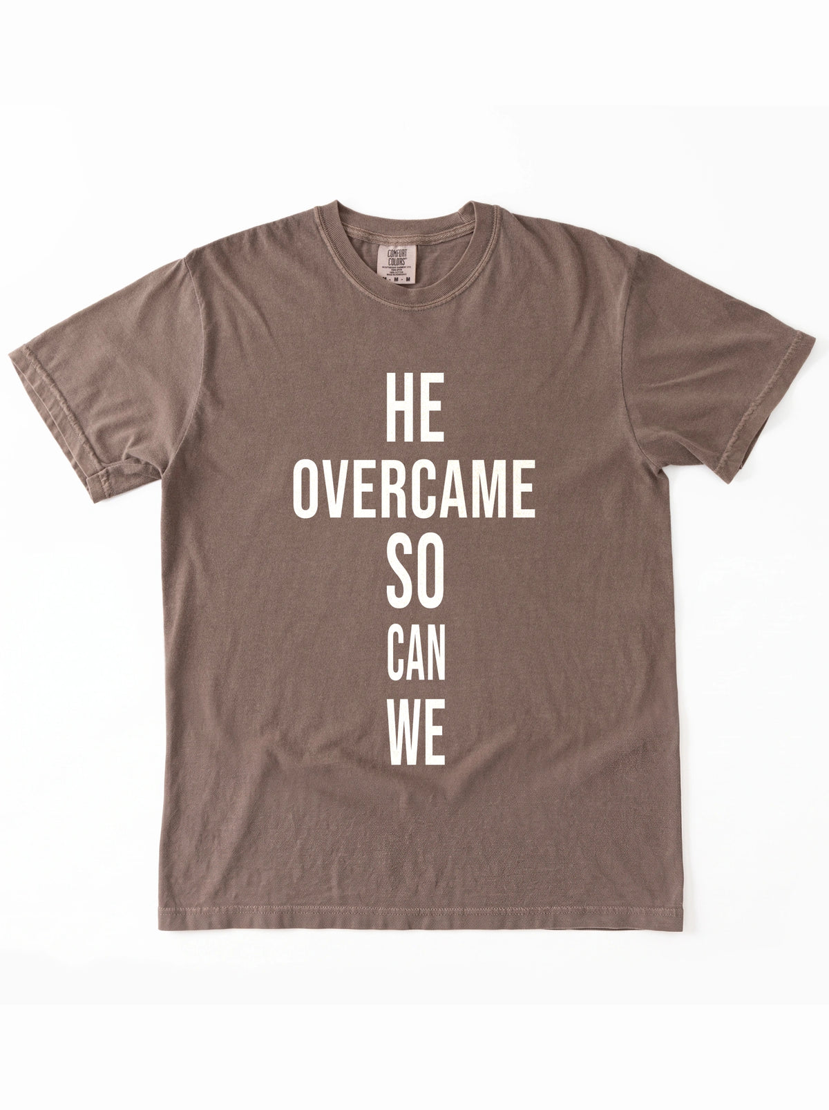 Faded brown T-shirt laid flat with large white text reading “HE OVERCAME SO CAN WE.”