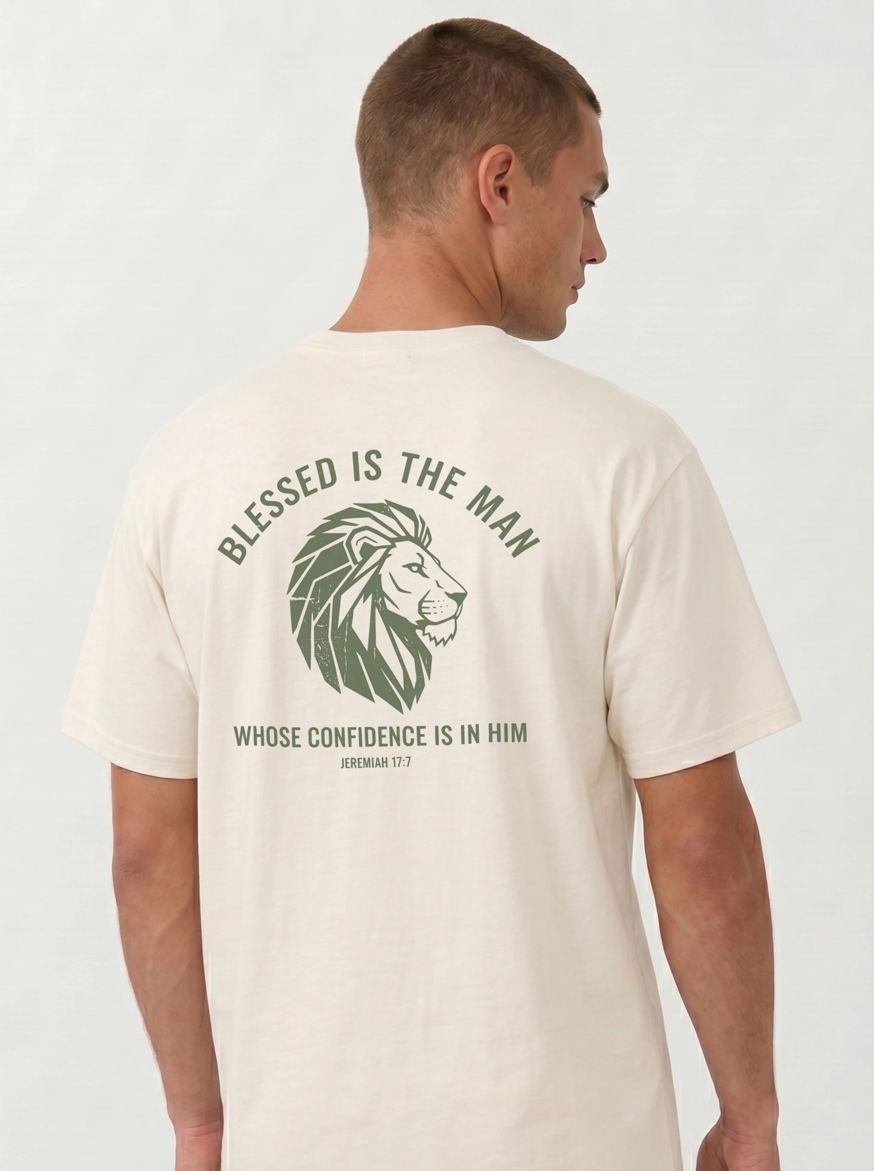 A man wearing a cream-colored t-shirt, viewed from the back. The shirt features a green graphic print of a lion's head with the text 'BLESSED IS THE MAN WHOSE CONFIDENCE IS IN HIM' and 'JEREMIAH 17:7'.
