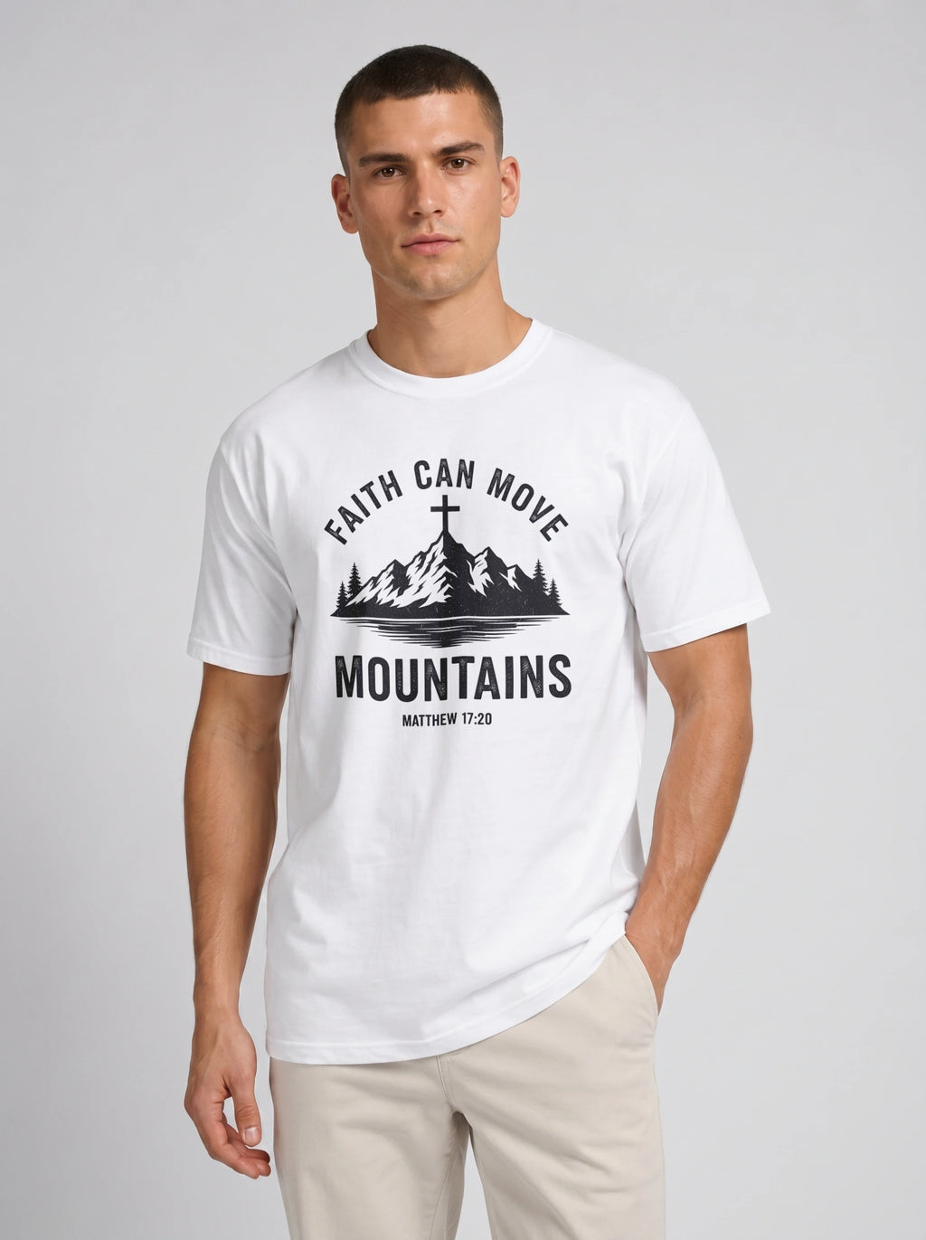 A man wearing a white short-sleeve graphic t-shirt with a black mountain and cross design and the text 'FAITH CAN MOVE MOUNTAINS MATTHEW 17:20', paired with beige pants, against a light gray studio background.