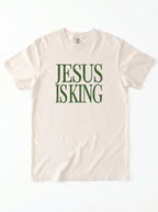 Cream T-shirt laid flat with distressed green text reading “Jesus Is King.”