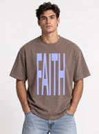 Person wearing a faded brown T-shirt with large light blue text reading “FAITH.”