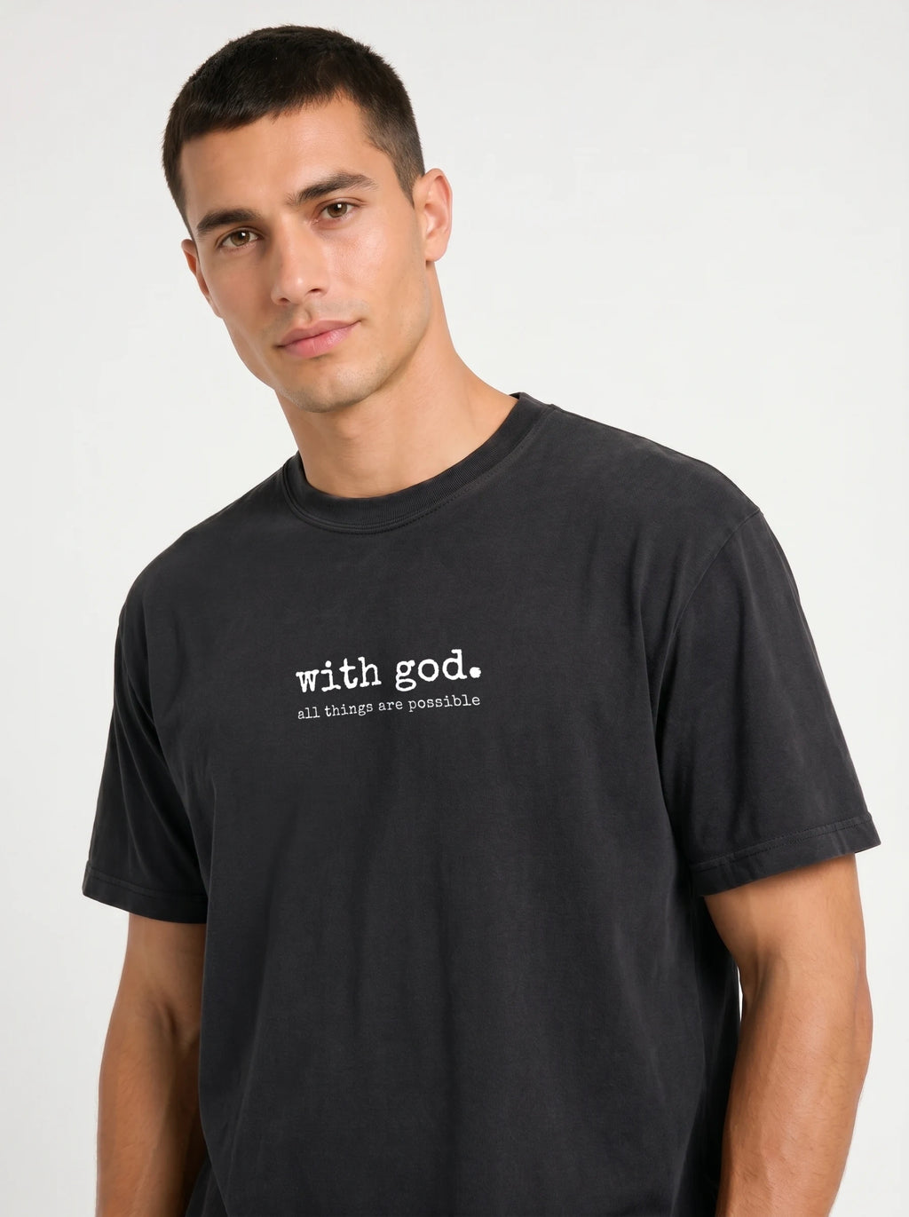 A man wearing a faded black crewneck t-shirt with the white printed text 'with god. all things are possible' on the chest, photographed against a light gray studio background.