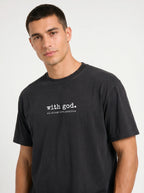 A man wearing a faded black crewneck t-shirt with the white printed text 'with god. all things are possible' on the chest, photographed against a light gray studio background.