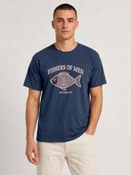 A man wearing a navy blue crewneck t-shirt with a graphic print of a fish and the text 'FISHERS OF MEN' and 'MATTHEW 4:19', paired with beige pants, against a light gray studio background.