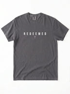 A dark gray crewneck t-shirt with the word 'REDEEMED' printed in white block letters across the chest, with a small white cross symbol underneath, displayed flat against a white background.