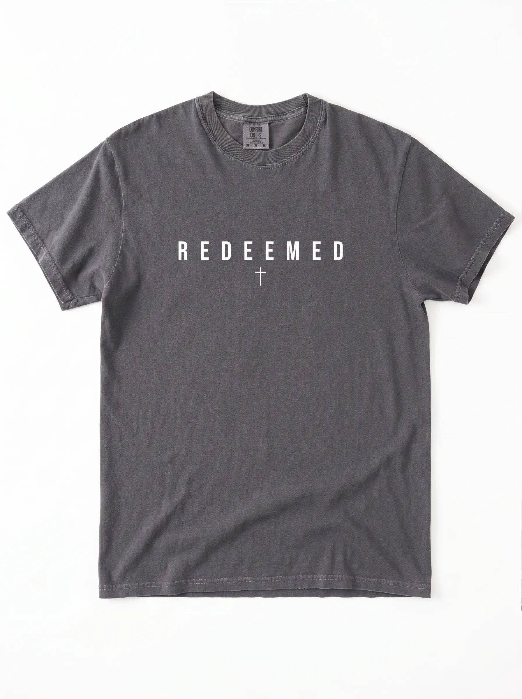 A dark gray crewneck t-shirt with the word 'REDEEMED' printed in white block letters across the chest, with a small white cross symbol underneath, displayed flat against a white background.