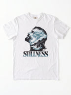 A white short-sleeve crewneck t-shirt featuring a graphic print of a man's profile filled with blue and white waves, with the word 'STILLNESS' printed below in a distressed blue font.
