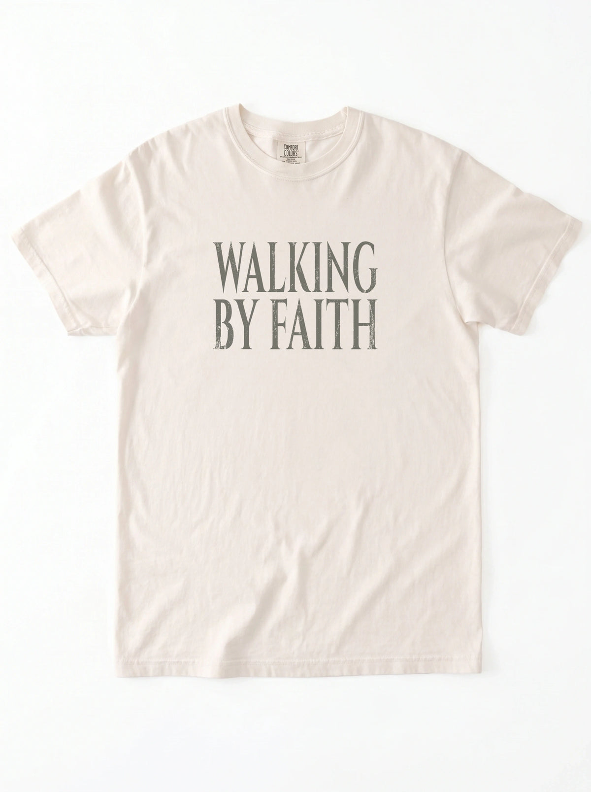 Cream T-shirt laid flat with distressed gray text reading “Walking By Faith.”