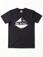 A black short-sleeve crewneck t-shirt with a white graphic on the chest showing a mountain peak, a lake reflection, and the text 'BE STILL' and 'PSALM 46:10'.