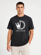 A man wearing a black short-sleeve t-shirt with a white graphic of a crown of thorns and the text 'LORD & SAVIOR' on the front, paired with light beige pants, against a plain light gray background.