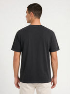 A man viewed from the back wearing a plain black short-sleeve crewneck t-shirt and light beige pants, set against a clean white studio background.