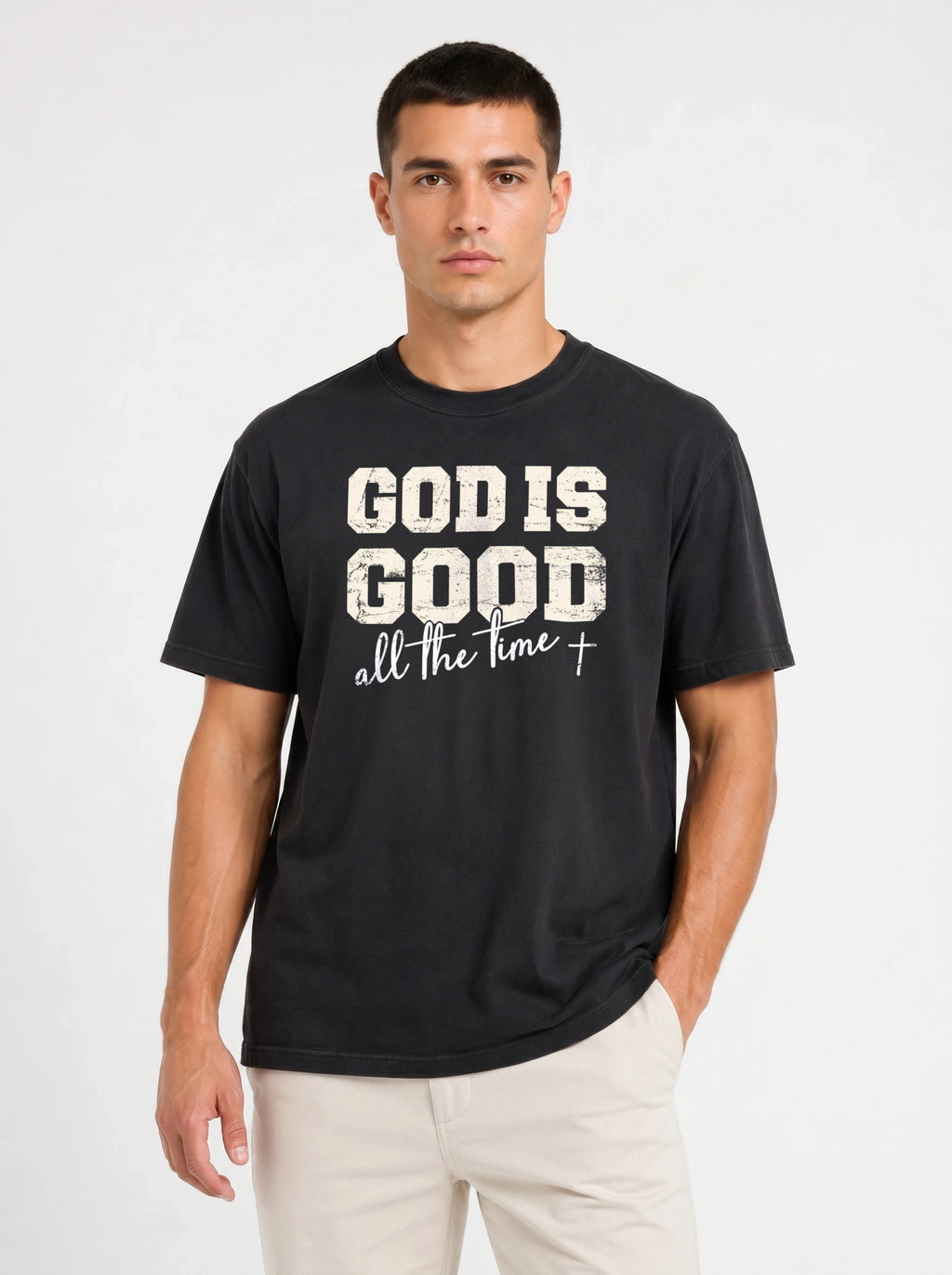 A man wearing a black crewneck t-shirt with a distressed white graphic that reads 'GOD IS GOOD all the time +', paired with light beige pants, against a plain light gray background.