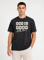 A man wearing a black crewneck t-shirt with a distressed white graphic that reads 'GOD IS GOOD all the time +', paired with light beige pants, against a plain light gray background.