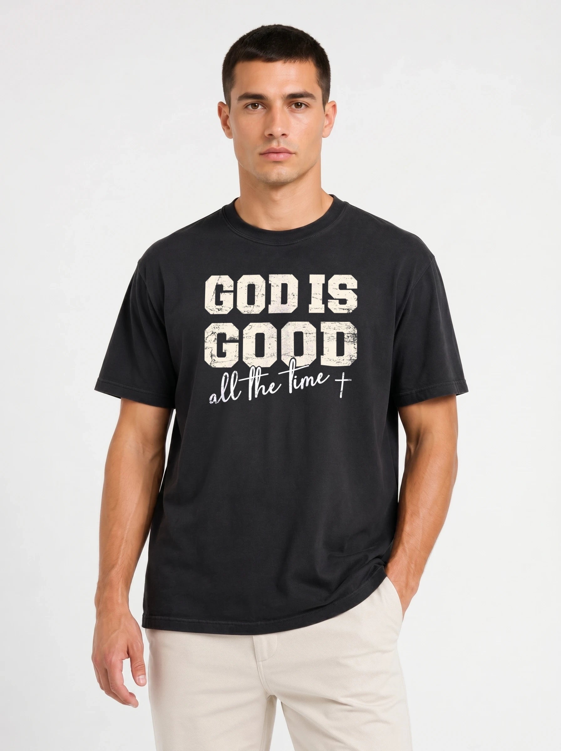 A man wearing a black crewneck t-shirt with a distressed white graphic that reads 'GOD IS GOOD all the time +', paired with light beige pants, against a plain light gray background.