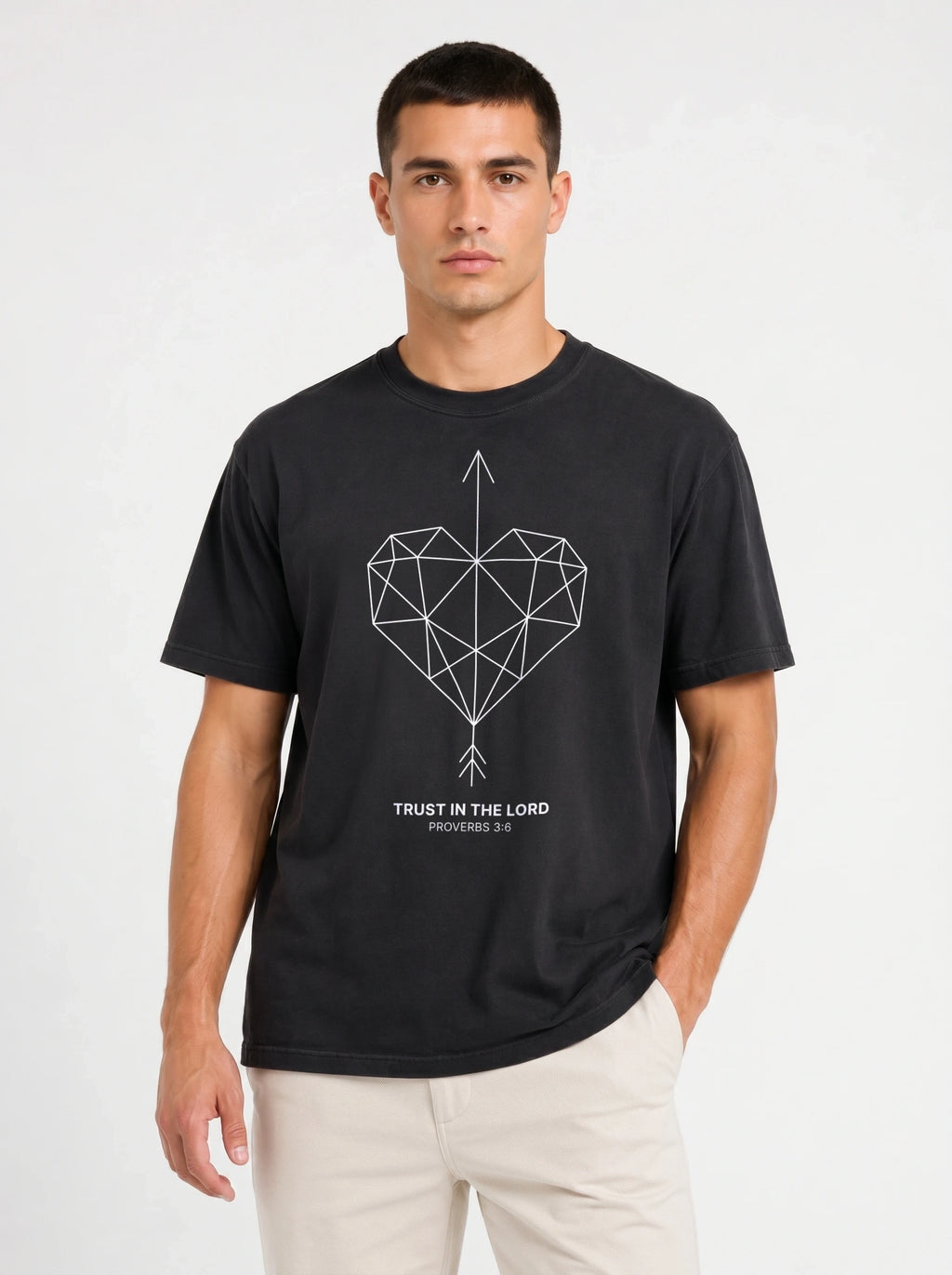 A man wearing a black short-sleeve t-shirt with a geometric heart pierced by an upward-pointing arrow, paired with the verse Proverbs 3:6: "Trust in the Lord.", paired with light beige pants, against a plain light gray background.