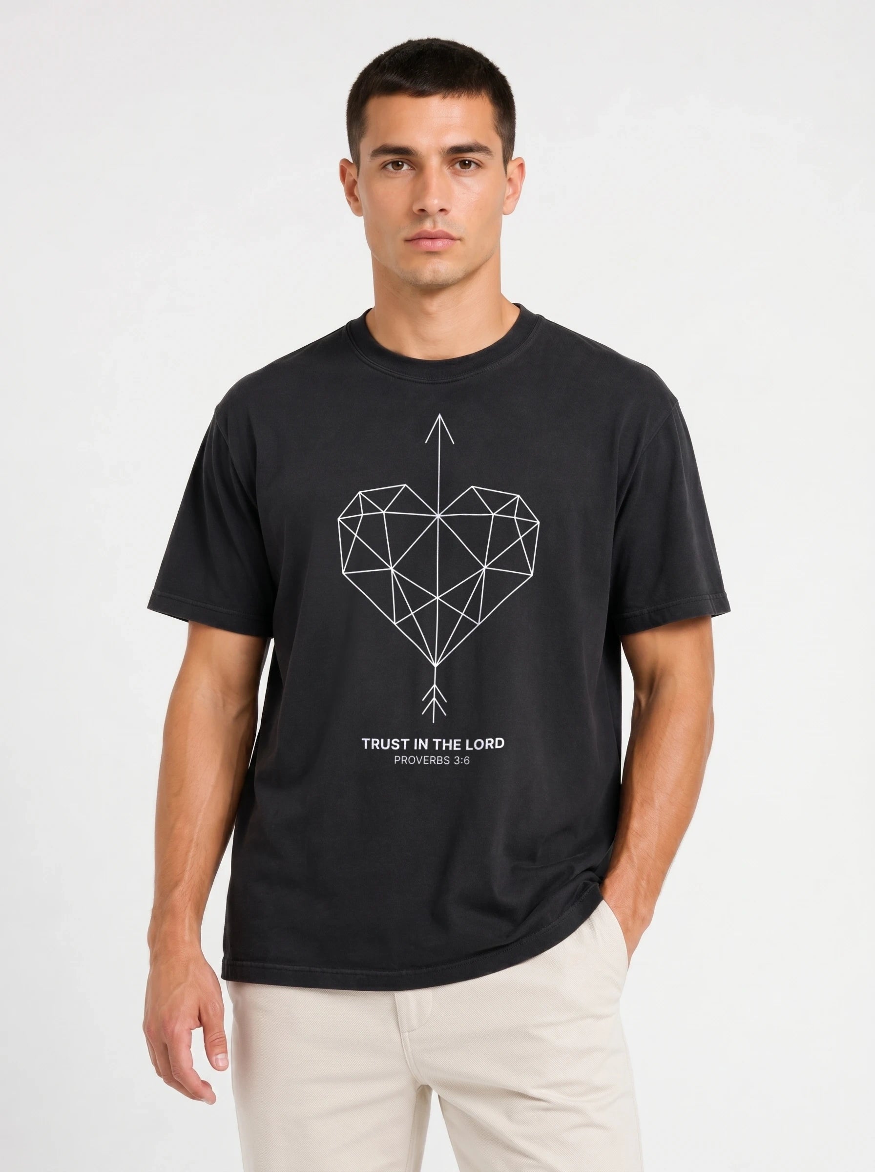 A man wearing a black short-sleeve t-shirt with a geometric heart pierced by an upward-pointing arrow, paired with the verse Proverbs 3:6: "Trust in the Lord.", paired with light beige pants, against a plain light gray background.