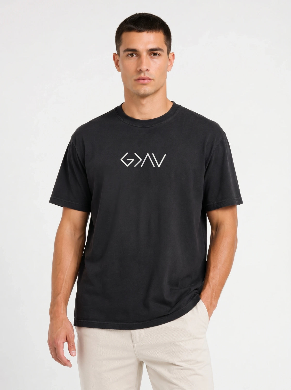 A man wearing a black short-sleeve t-shirt with a white stylized 'God Is Greater Than Highs and Lows' logo on the chest, paired with light beige pants, against a plain light gray background.