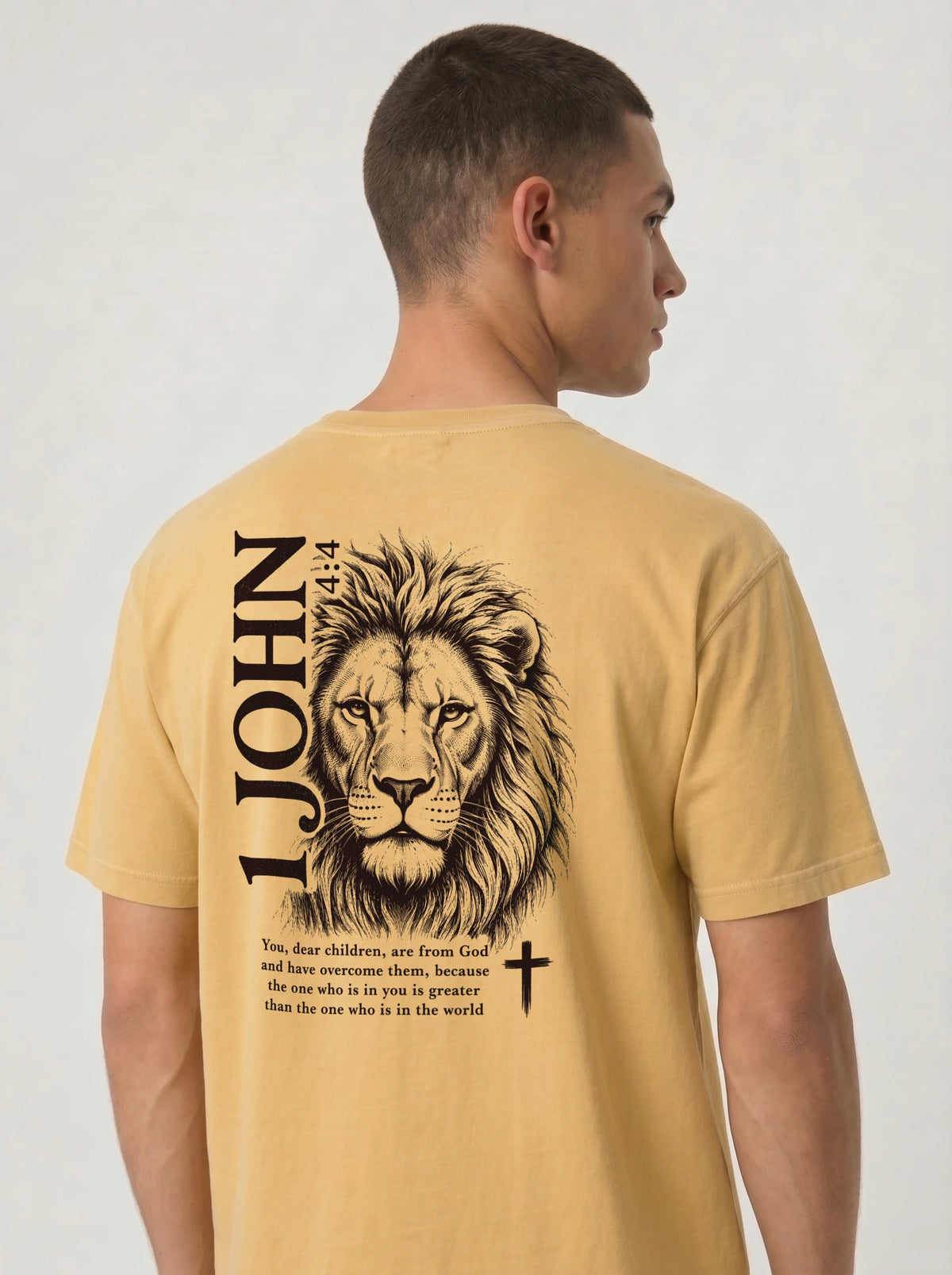 Person wearing a yellow T-shirt with a black lion illustration, cross, “1 John 4:4,” and the verse text, “You, dear children, are from God and have overcome them, because the one who is in you is greater than the one who is in the world.”