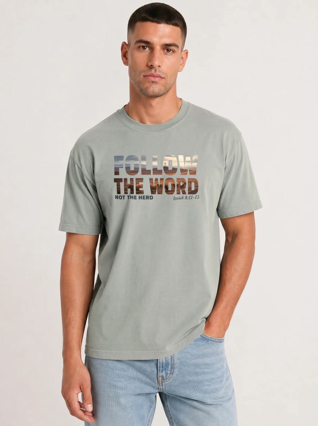 A man wearing a light sage green graphic t-shirt with the text 'FOLLOW THE WORD NOT THE HERD' and 'Isaiah 8:11-13' in a textured, multi-toned font printed on the chest, paired with light blue jeans, against a plain light gray background.