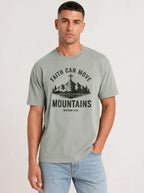 A man wearing a sage green graphic t-shirt with a black mountain and cross design that reads 'FAITH CAN MOVE MOUNTAINS MATTHEW 17:20' with a mountain and cross illustration, paired with light blue jeans, against a plain light gray background.