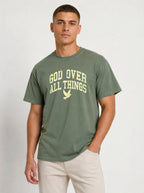 Medium front-facing shot of a male model wearing a muted sage green short-sleeve t-shirt and light beige shorts, featuring a distressed yellow "GOD OVER ALL THINGS" graphic and dove icon.