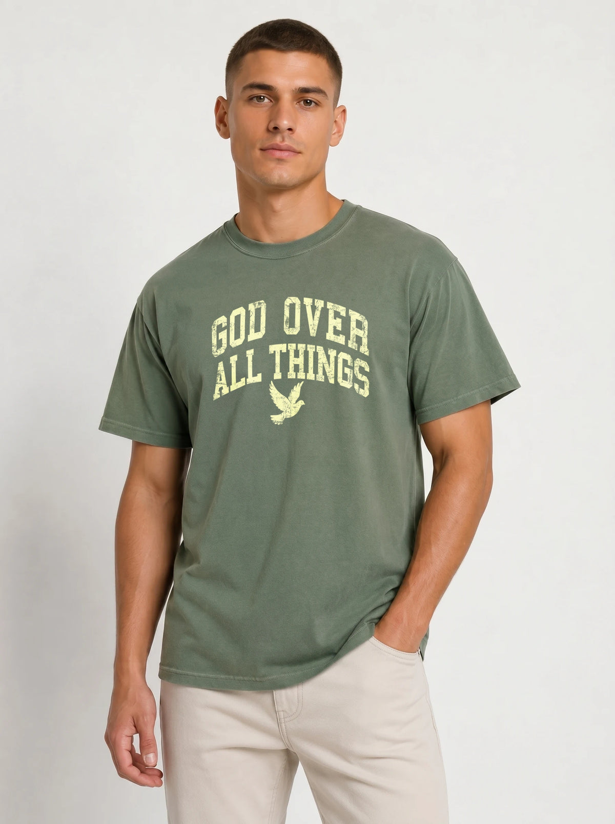 Medium front-facing shot of a male model wearing a muted sage green short-sleeve t-shirt and light beige shorts, featuring a distressed yellow "GOD OVER ALL THINGS" graphic and dove icon.