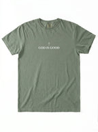 Muted green T-shirt laid flat with a small cross above the text “God Is Good.”