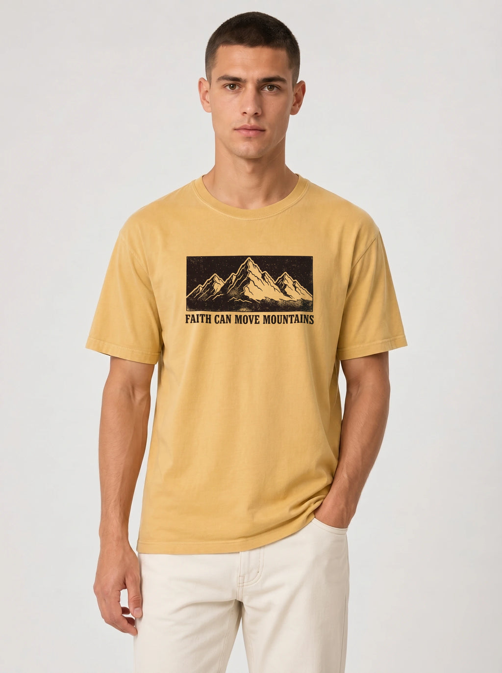 A man wearing a mustard yellow t-shirt with a black graphic of mountains and the text 'FAITH CAN MOVE MOUNTAINS' on the chest, paired with off-white pants, against a light gray background.