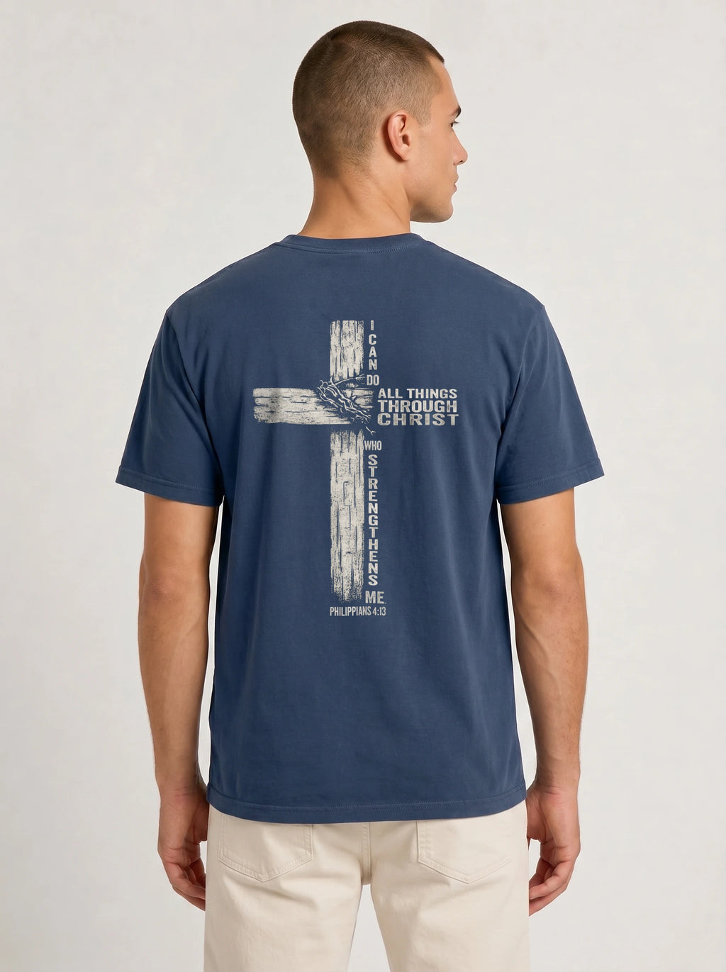 A man viewed from the back wearing a navy blue t-shirt with a distressed biege graphic of a cross and the text 'I CAN DO ALL THINGS THROUGH CHRIST WHO STRENGTHENS ME' from Philippians 4:13, paired with beige pants against a light gray studio background.
