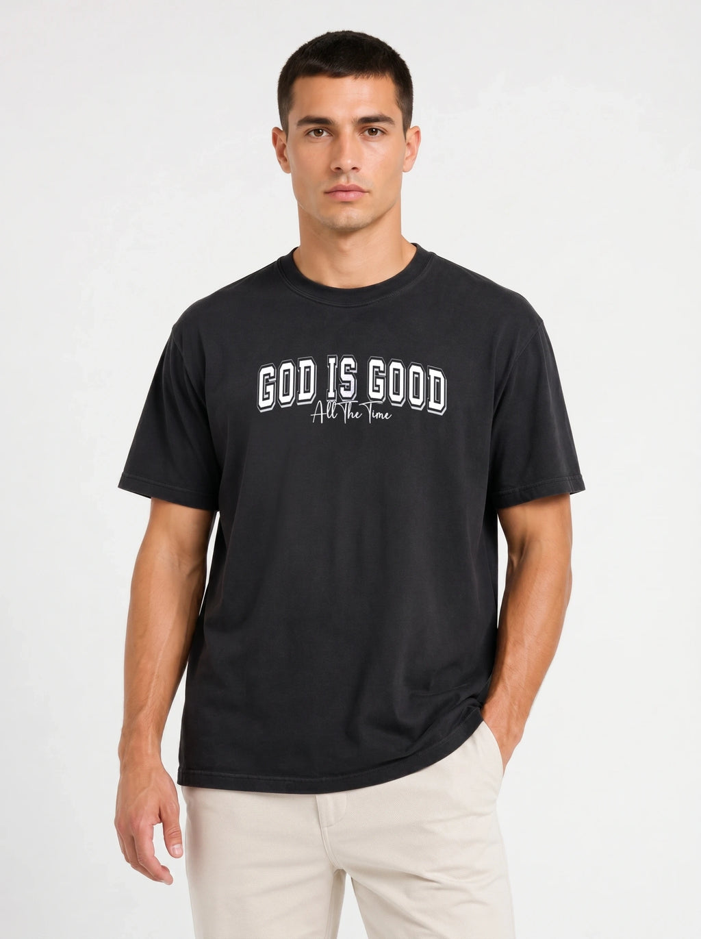 A man wearing a black short-sleeve t-shirt with the text 'GOD IS GOOD All The Time' printed in white on the chest, paired with light beige pants, against a plain light gray background.
