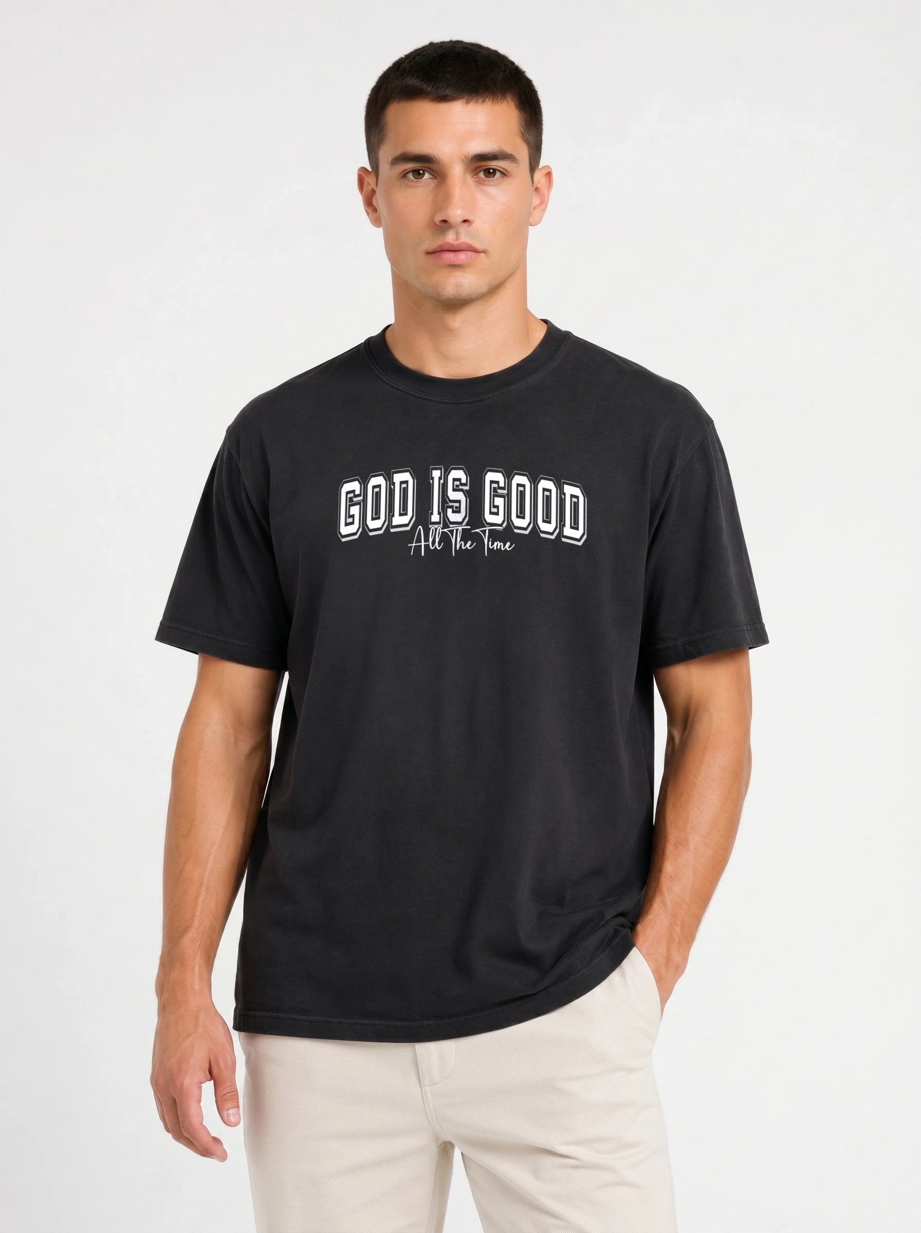 A man wearing a black short-sleeve t-shirt with the text 'GOD IS GOOD All The Time' printed in white on the chest, paired with light beige pants, against a plain light gray background.