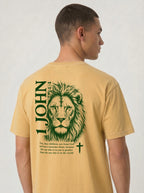 Person wearing a faded yellow T-shirt with a green lion illustration, cross, “1 John 4:4,” and the verse text, “You, dear children, are from God and have overcome them, because the one who is in you is greater than the one who is in the world.”