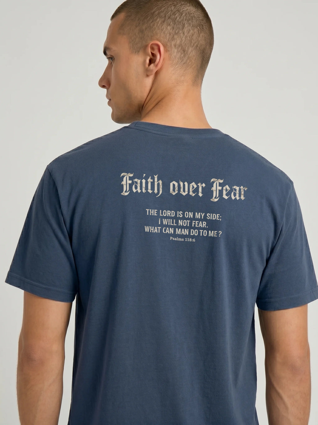 A man wearing a navy blue t-shirt, viewed from the back. The shirt features the text 'Faith over Fear' in a gothic font, followed by a bible verse from Psalms 118:6 in biege print.