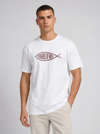 A man wearing a white crewneck t-shirt with a dark red graphic logo on the chest that reads 'FISHERS OF MEN' inside a fish shape, paired with beige pants, against a light gray studio background.