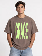 Person wearing a faded brown T-shirt with large neon green text reading “GRACE.”