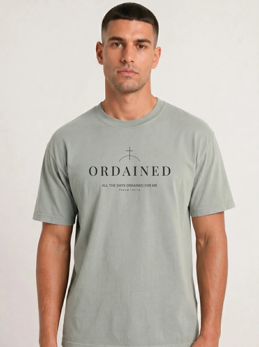 A man wearing a light sage green crewneck t-shirt with a black printed graphic on the chest that reads 'ORDAINED' and 'ALL THE DAYS ORDAINED FOR ME' below it, set against a plain light gray background.