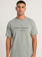 A man wearing a light sage green crewneck t-shirt with a black printed graphic on the chest that reads 'ORDAINED' and 'ALL THE DAYS ORDAINED FOR ME' below it, set against a plain light gray background.