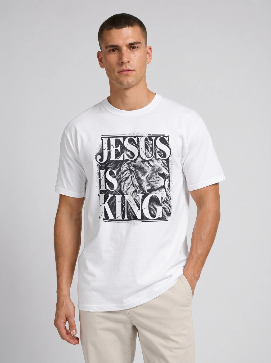 A man wearing a white crewneck t-shirt with a black graphic print that reads 'JESUS IS KING' alongside an illustration of a lion's head, paired with beige pants, against a light gray studio background.