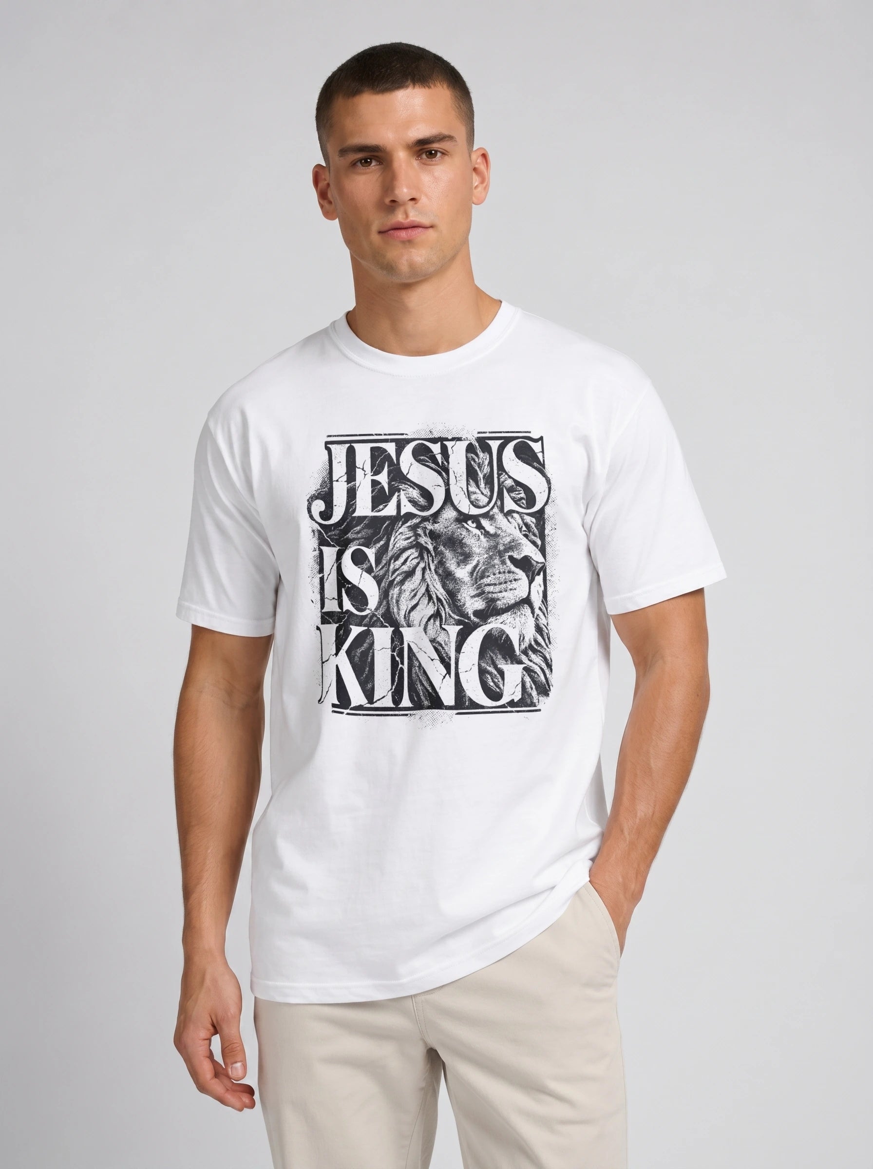 A man wearing a white crewneck t-shirt with a black graphic print that reads 'JESUS IS KING' alongside an illustration of a lion's head, paired with beige pants, against a light gray studio background.