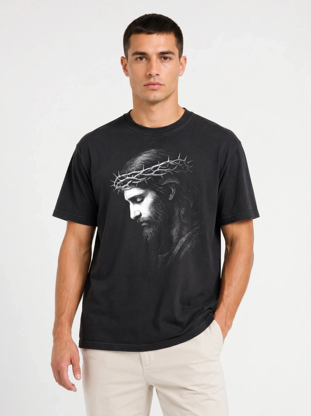 A man wearing a black crew-neck t-shirt with a grayscale graphic print of Jesus Christ wearing a crown of thorns, paired with off-white trousers, against a plain light gray background.