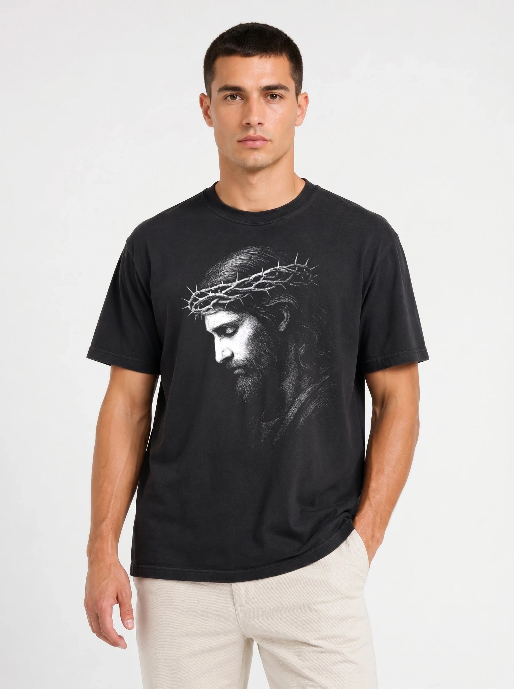 A man wearing a black crew-neck t-shirt with a grayscale graphic print of Jesus Christ wearing a crown of thorns, paired with off-white trousers, against a plain light gray background.