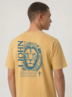 A man wearing a mustard yellow garment-dyed t-shirt, viewed from an angled back view. The shirt features a large blue lion illustration with '1 JOHN 4:4' text, a Bible verse, and a small cross symbol.