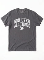 Close-up portrait of a male model looking forward, wearing a charcoal t-shirt featuring a distressed white graphic with the words "GOD OVER ALL THINGS" and a flying dove icon.